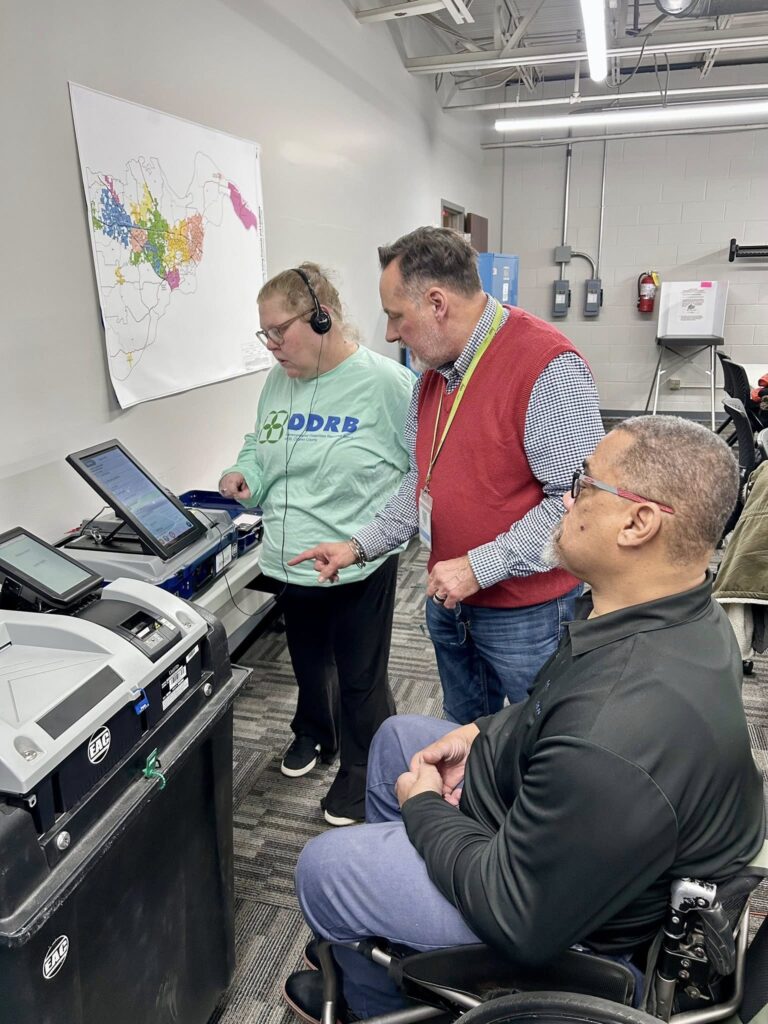 Three people gather around voting machines to discuss accessible voting practices.